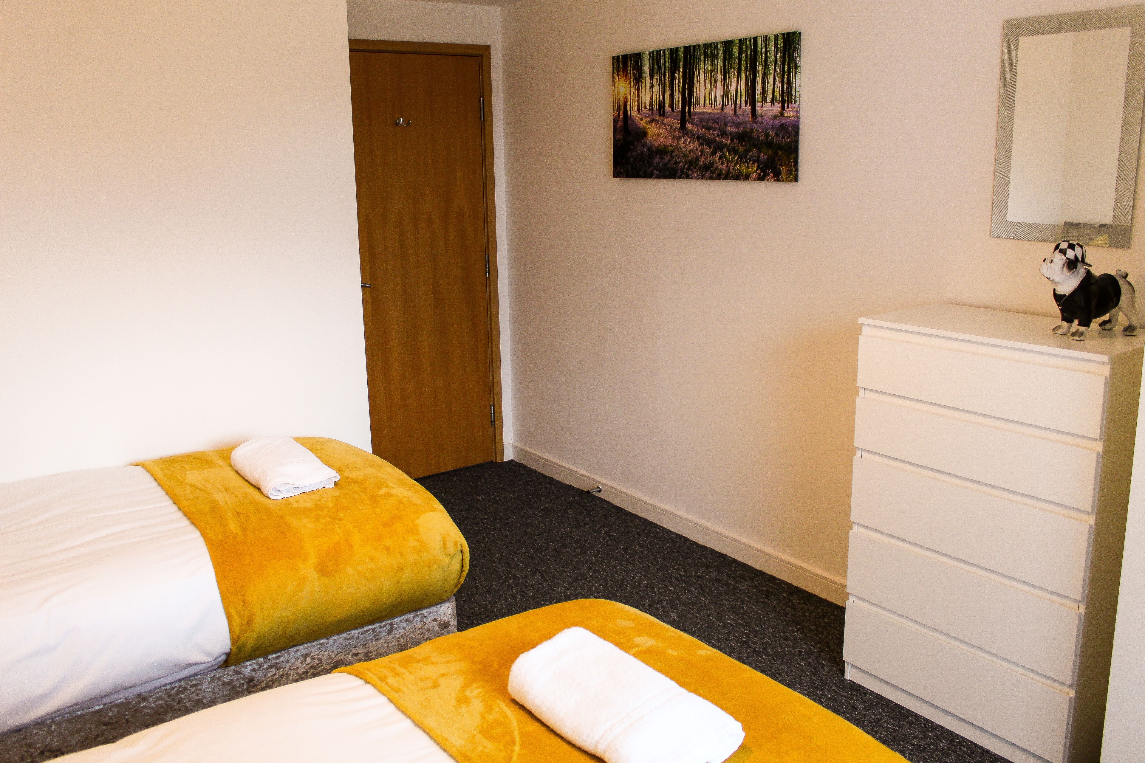 A hotel room with two beds, a wooden door, a painting, a dresser, and a small dog figurine.