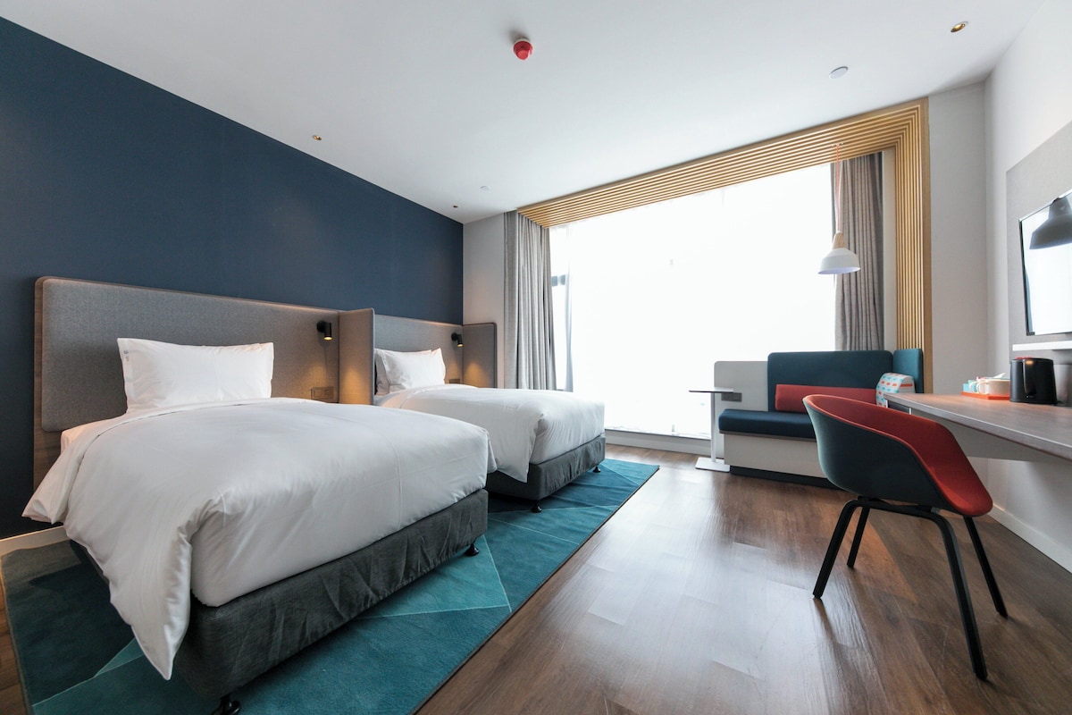 Ett hotellrum med två sängar, ett skrivbord, en stol och ett stort fönster.
