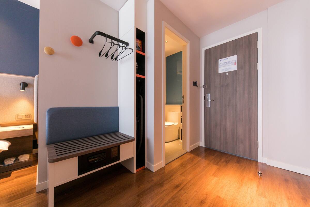 A modern hotel room with a wooden floor, a bench, a coat rack, and a door leading to a bathroom.