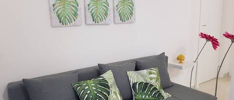 Un canapé gris agrémenté de deux coussins à motifs verts et blancs, un mur blanc orné de quatre œuvres d’art représentant des feuilles encadrées, et une petite étagère blanche sur laquelle repose un objet jaune.