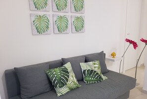 Un divano grigio con due cuscini verde e bianchi a motivi, una parete bianca con quattro opere d'arte raffiguranti foglie e un piccolo scaffale con un oggetto giallo.
