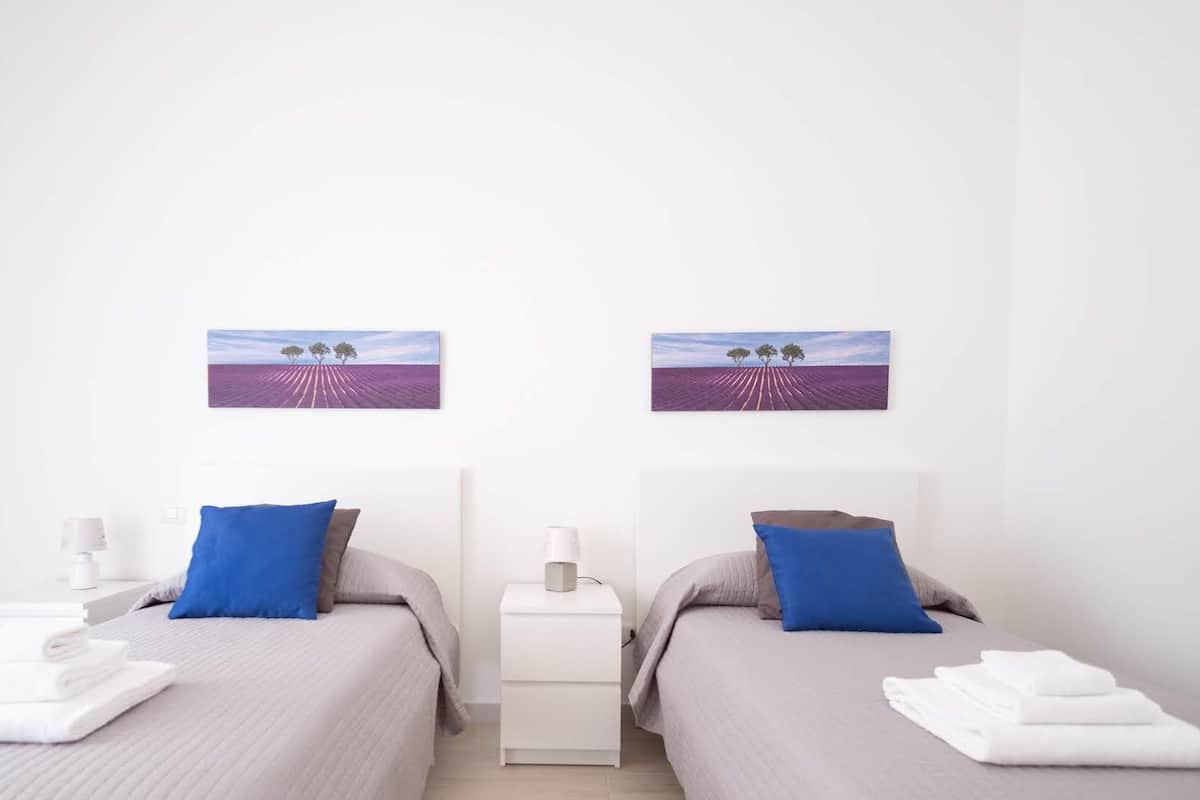 A hotel room with two beds, each with a blue pillow and a white towel. There are two nightstands with lamps and a small table between the beds.