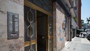 A building facade with a metal nameplate, a large glass door with a geometric pattern, and a sidewalk with parked cars and pedestrians. - Casa Viviana - Bari Policlinico (Bari)