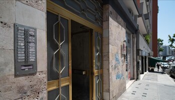 Facciata di un edificio con una targa metallica, un'ampia porta in vetro con motivo geometrico e un marciapiede con auto parcheggiate e pedoni.