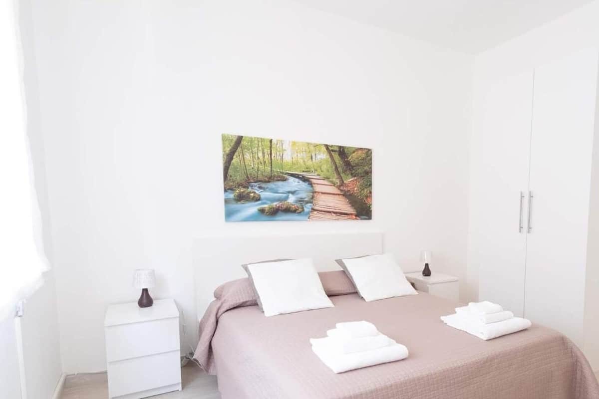 A bedroom with a bed, white nightstands, a wardrobe, and a painting of a forest stream.