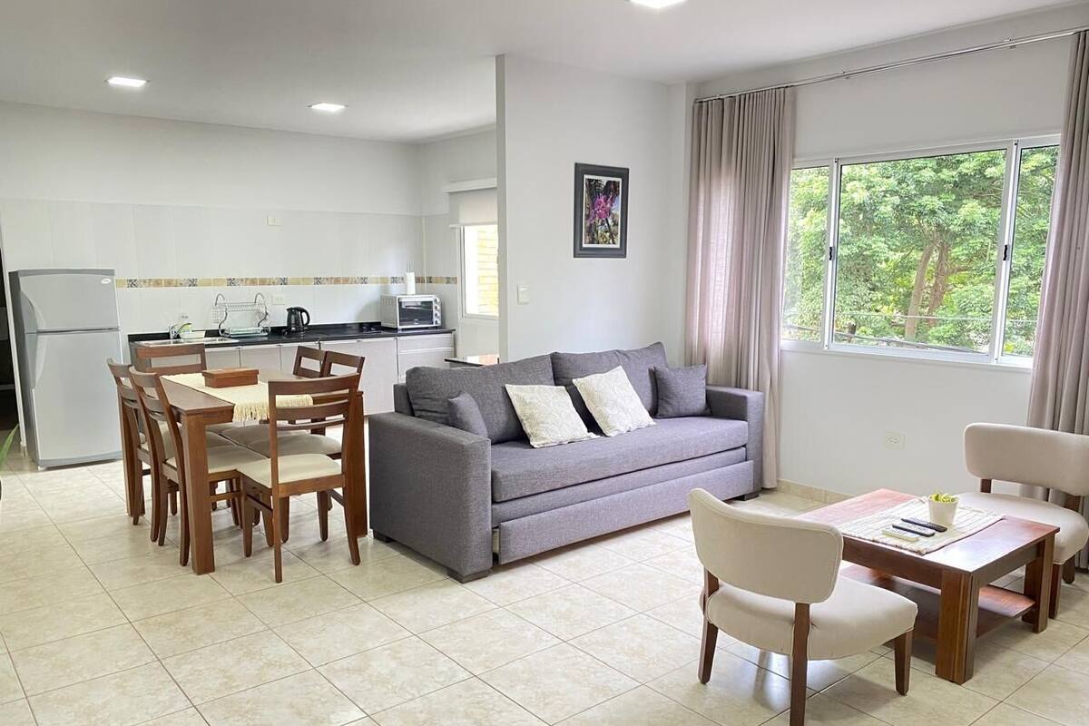 A modern living room with a grey sofa, wooden dining table, and chairs. A kitchen with white cabinets and a refrigerator is visible in the background.