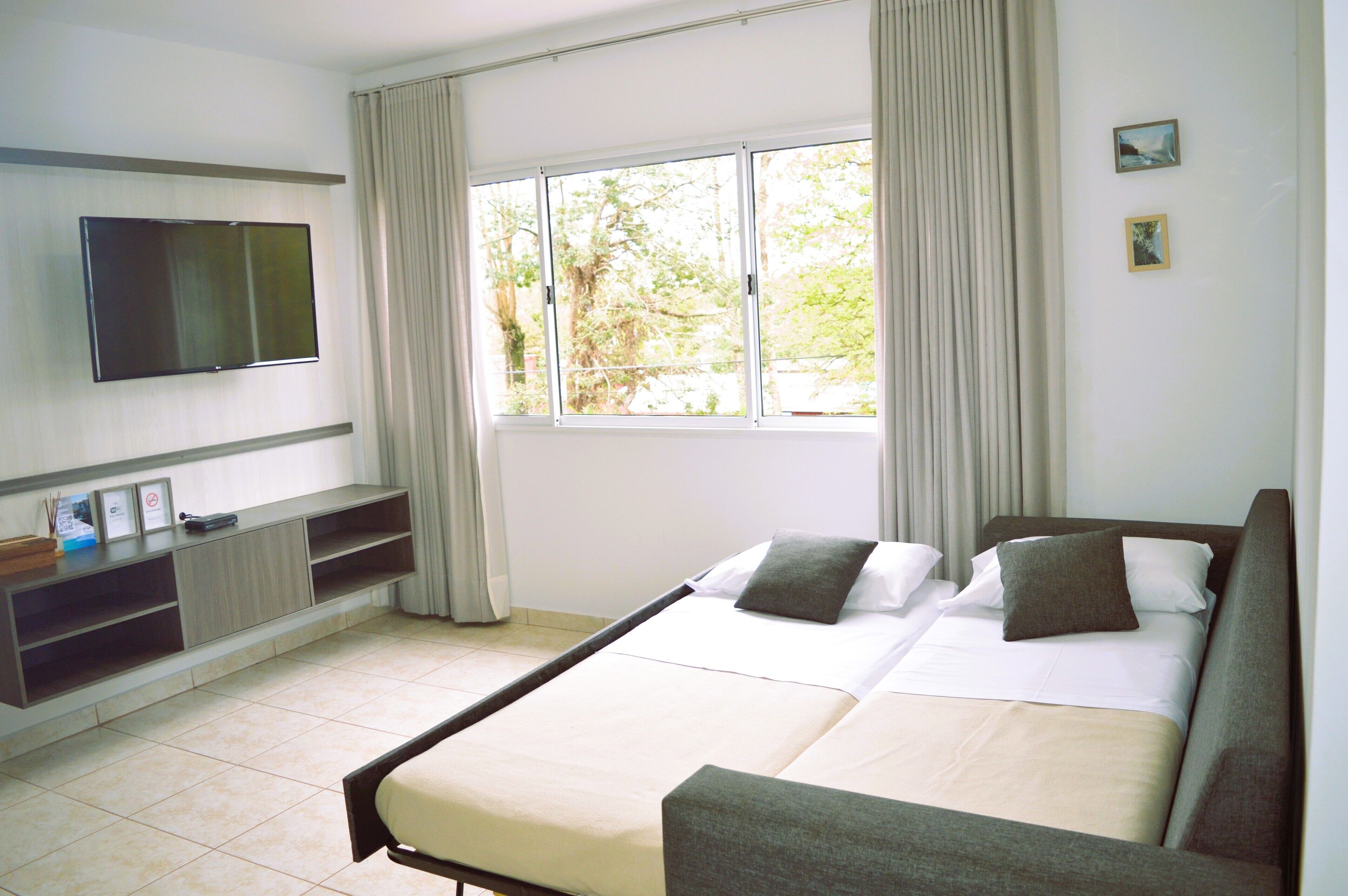 Habitación de hotel con cama, televisor montado en la pared, ventana con cortinas y una pequeña estantería con objetos.