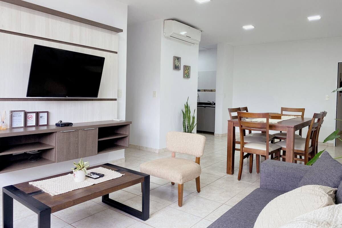 A modern living room with a flat-screen TV, wooden furniture, a dining area, and a kitchen in the background.