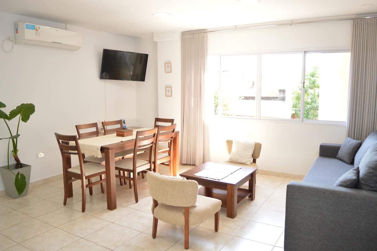 A living room with a dining area, a grey sofa, a TV mounted on the wall, and a large window with curtains.