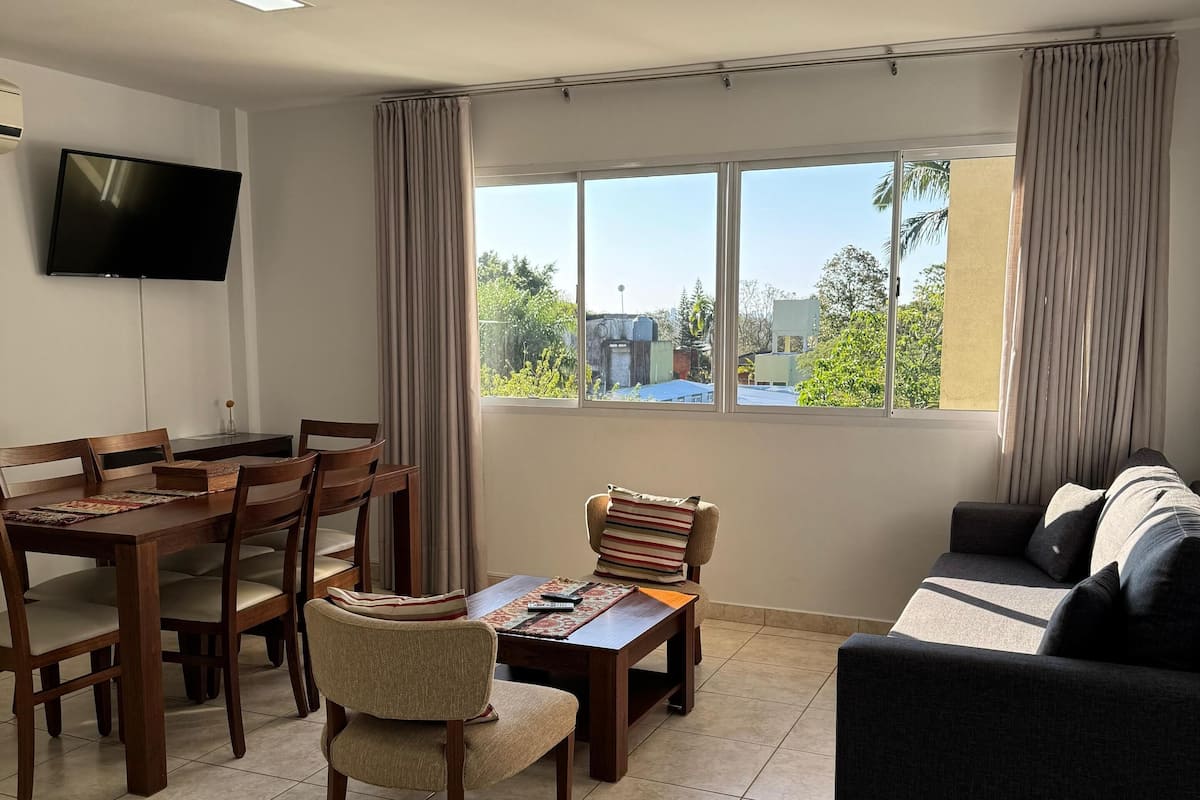 A living room with a dining area, a flat-screen TV mounted on the wall, a large window with curtains, and a sofa.
