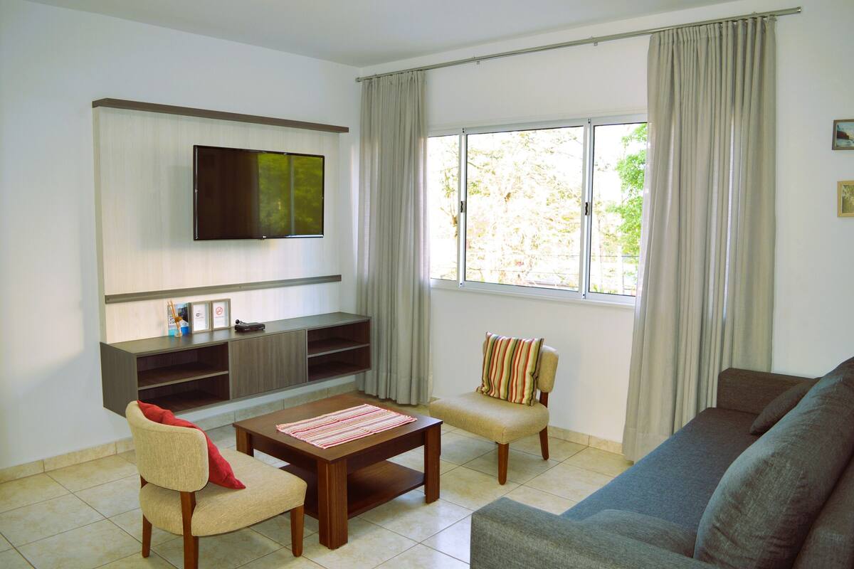A living room with a flat-screen TV mounted on the wall, a sofa, two armchairs, a coffee table, and a sliding glass door leading to a balcony.