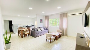 Una sala de estar moderna con un sofá gris, una mesa de comedor de madera y sillas. Hay una zona de cocina con un refrigerador y microondas. Un televisor está montado en la pared, y se ve una ventana con cortinas.