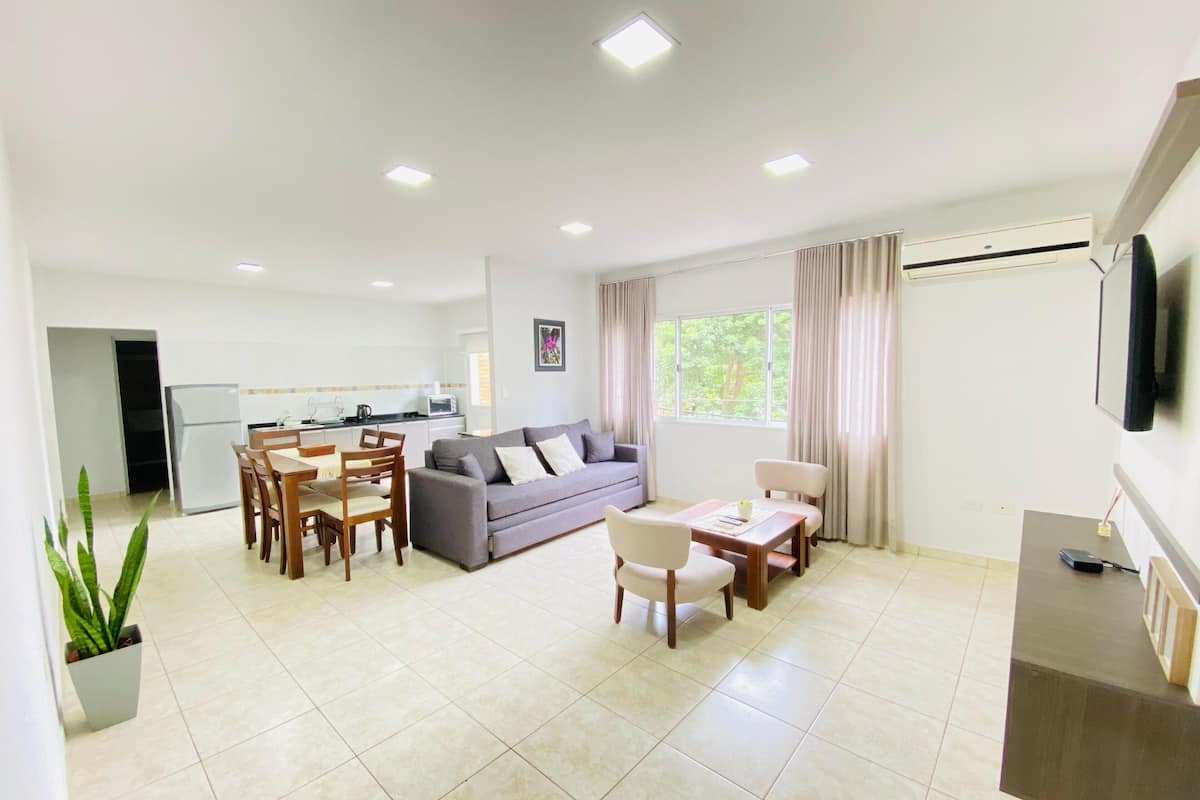 A modern living room with a grey sofa, wooden dining table, and chairs. There is a kitchen area with a refrigerator and microwave. A TV is mounted on the wall, and a window with curtains is visible.