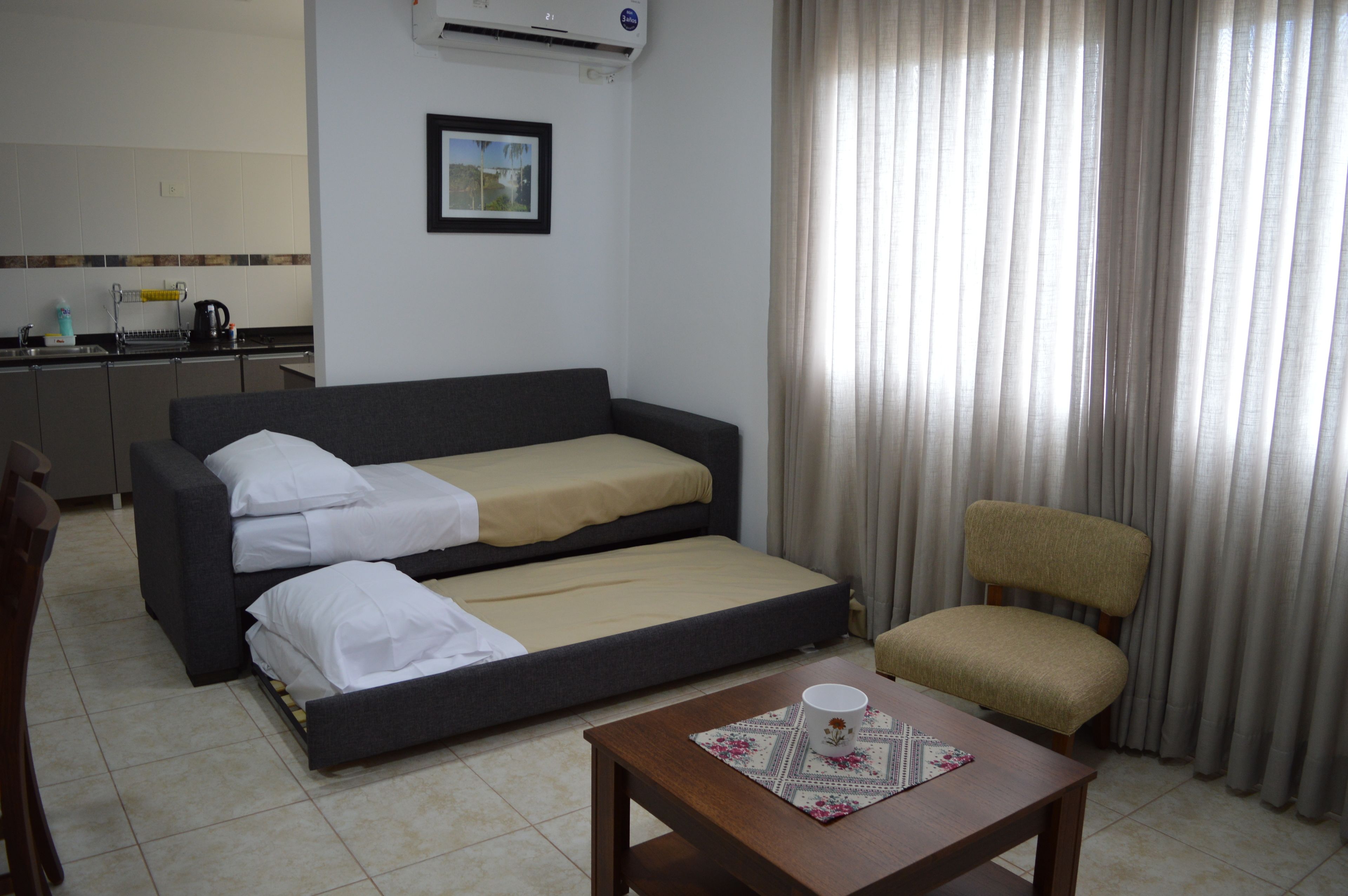 Ein kompaktes Hotelzimmer mit Schlafsofa, einem kleinen Holztisch, einem Stuhl und einer Küchenzeile im Hintergrund.