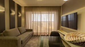 A modern living room with a sofa, a glass coffee table, a flat-screen TV mounted on the wall, and a large window with sheer curtains. - Hotel Home Vacations (Natal)