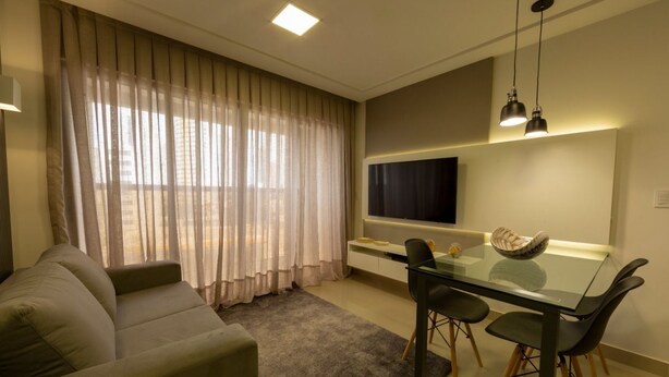 A modern living room with a sofa, a glass dining table with chairs, a flat-screen TV mounted on the wall, and sheer curtains on the windows. - Hotel Home Vacations (Natal)