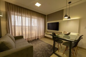 A modern living room with a sofa, a glass dining table with chairs, a flat-screen TV mounted on the wall, and sheer curtains on the windows. - Hotel Home Vacations (Natal)