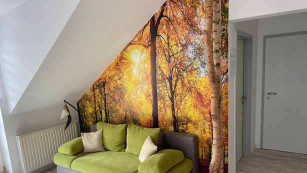 A modern living room with a green sofa, a large forest-themed tapestry, and a white door. - Hotel Garni Zum Holzfäller (Wernigerode)