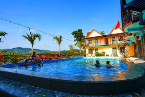 A swimming pool with people, a building with a red roof, and palm trees in the background.