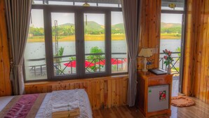 A room with a bed, a small desk, a refrigerator, and a large window overlooking a body of water and a dock with umbrellas. - Phong Nha Escape Bungalow (Phong Nha)