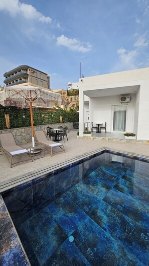 A swimming pool with lounge chairs and tables, a white building with a door and window, and a building with balconies in the background. - Vila Finnley (Ksamil)