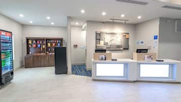 A hotel reception area with a Coca-Cola refrigerator, snack vending machine, and a reception desk with a digital screen.