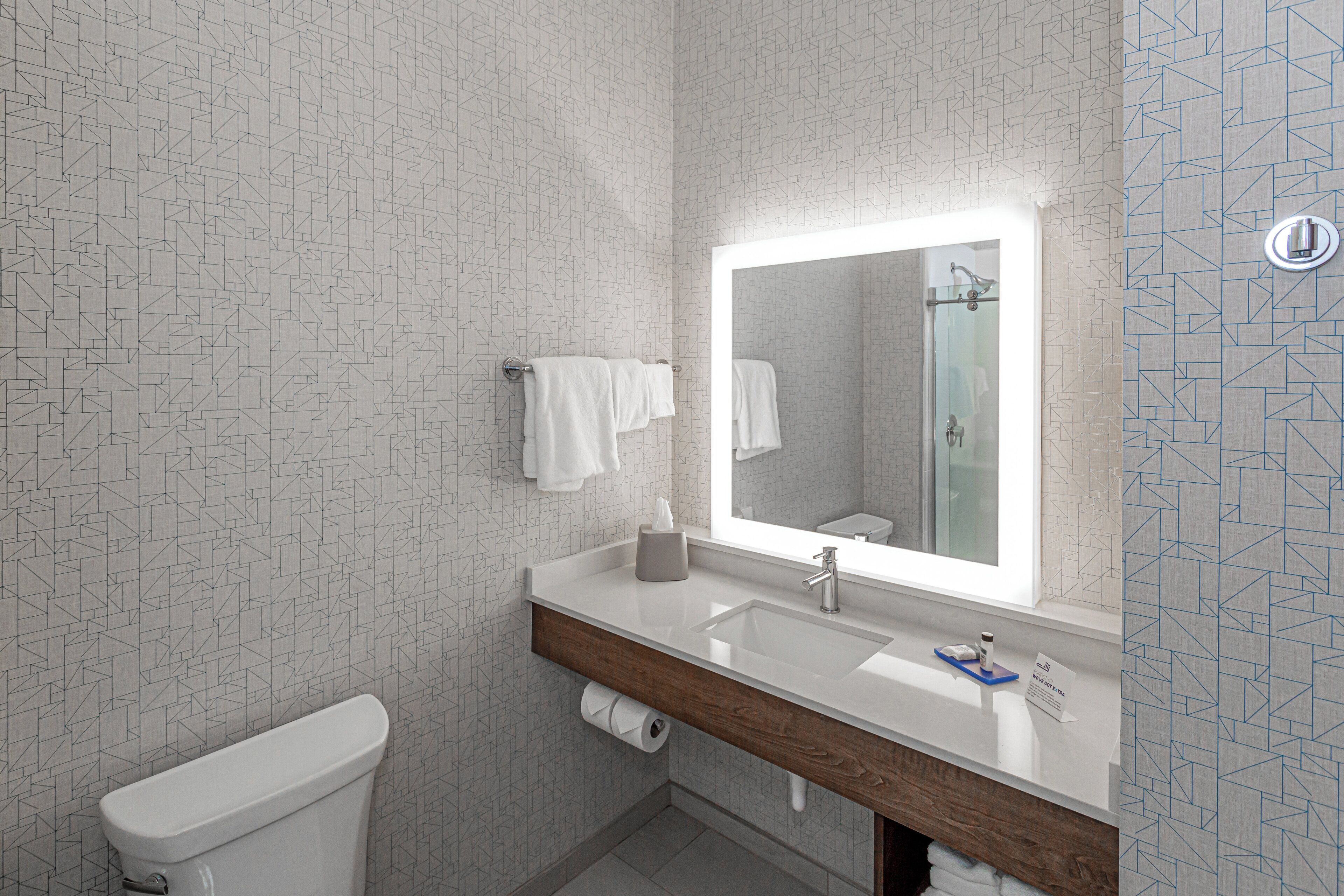 A modern bathroom with a geometric patterned wall, a white toilet, a sink with a mirror, and a towel rack.