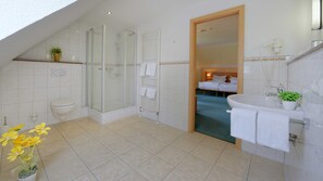 Ein modernes Badezimmer mit Dusche, Toilette und Waschbecken. Im Hintergrund ist ein Bett durch eine geöffnete Tür zu sehen.
