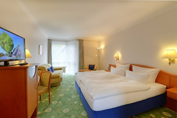 A hotel room with a large bed, a desk with a television, a chair, a small table, and a lamp.