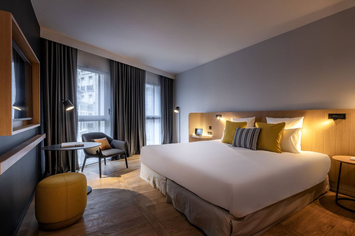 Une chambre d’hôtel moderne avec un grand lit, une petite table ronde, un canapé et un pouf jaune.