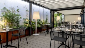 Un área de comedor al aire libre con mesas y sillas, rodeada de plantas y una pérgola.