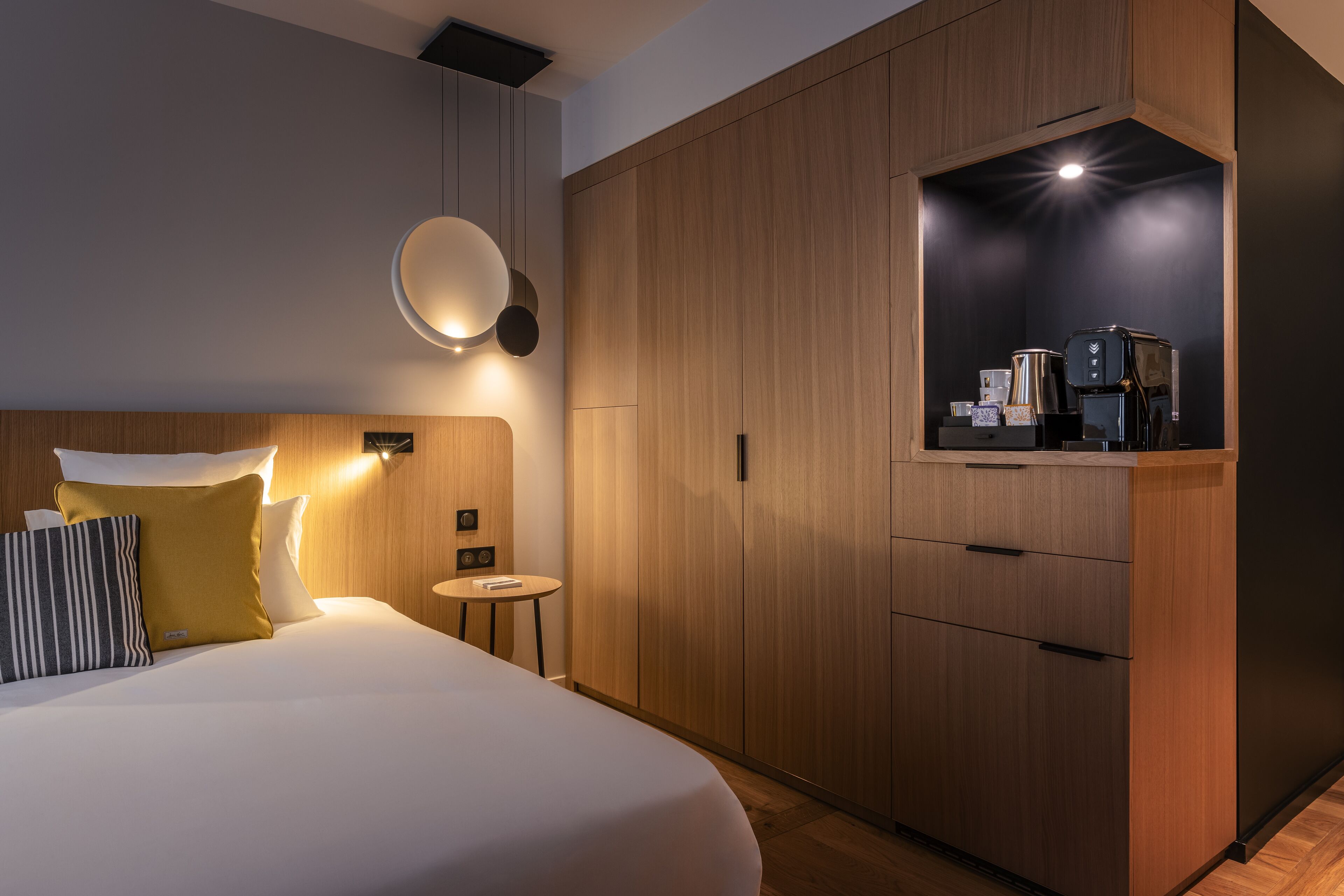 A modern hotel room with a large bed, bedside table, and a built-in kitchenette with a coffee maker.