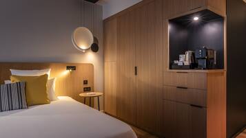 A modern hotel room with a large bed, bedside table, and a built-in kitchenette with a coffee maker.