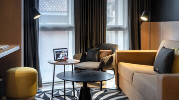 Una habitación de hotel moderna con sofá, sillón, reposapiés y una mesita con un marco de fotos.