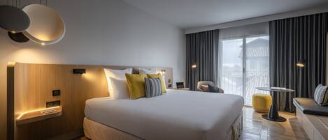 Una habitación de hotel moderna con una cama grande, mesitas de noche, un área de estar con sofá y sillón, y vistas de la ciudad a través de amplias ventanas.