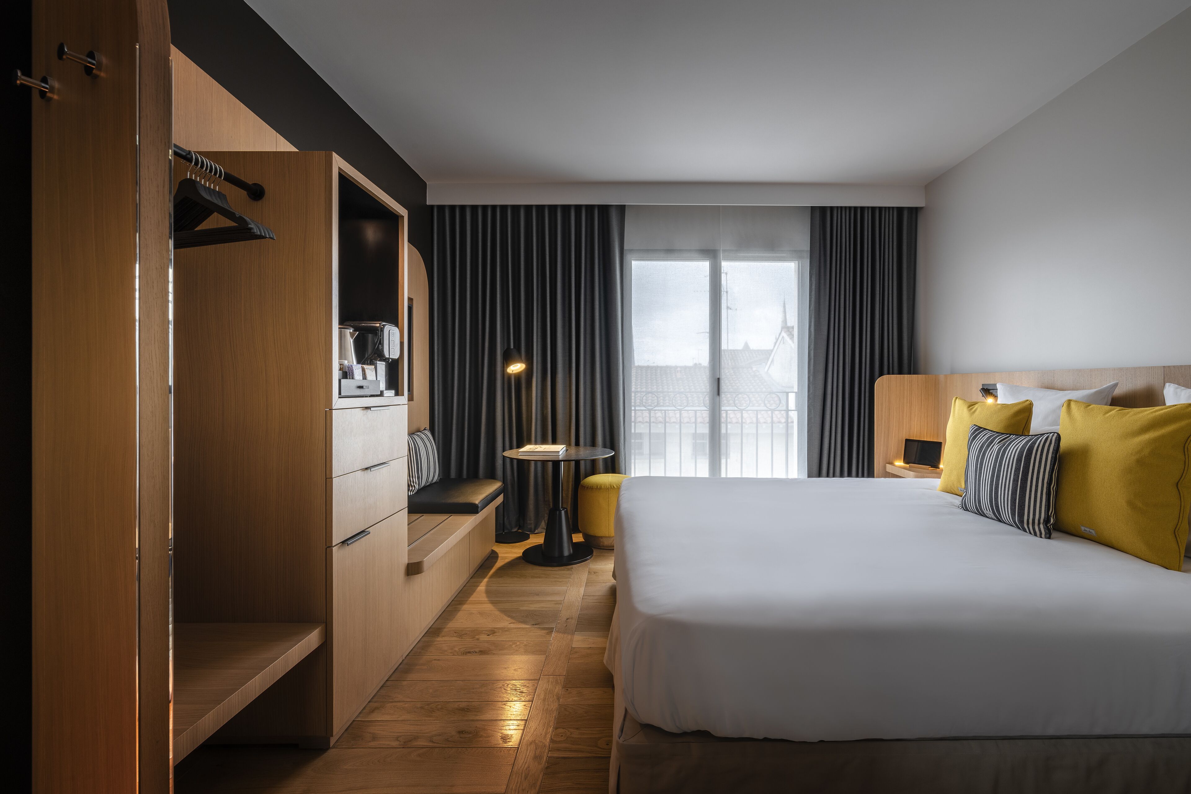 Habitación de hotel con una cama grande, un armario de madera, una mesita con lámpara y vistas a la ciudad desde la ventana.