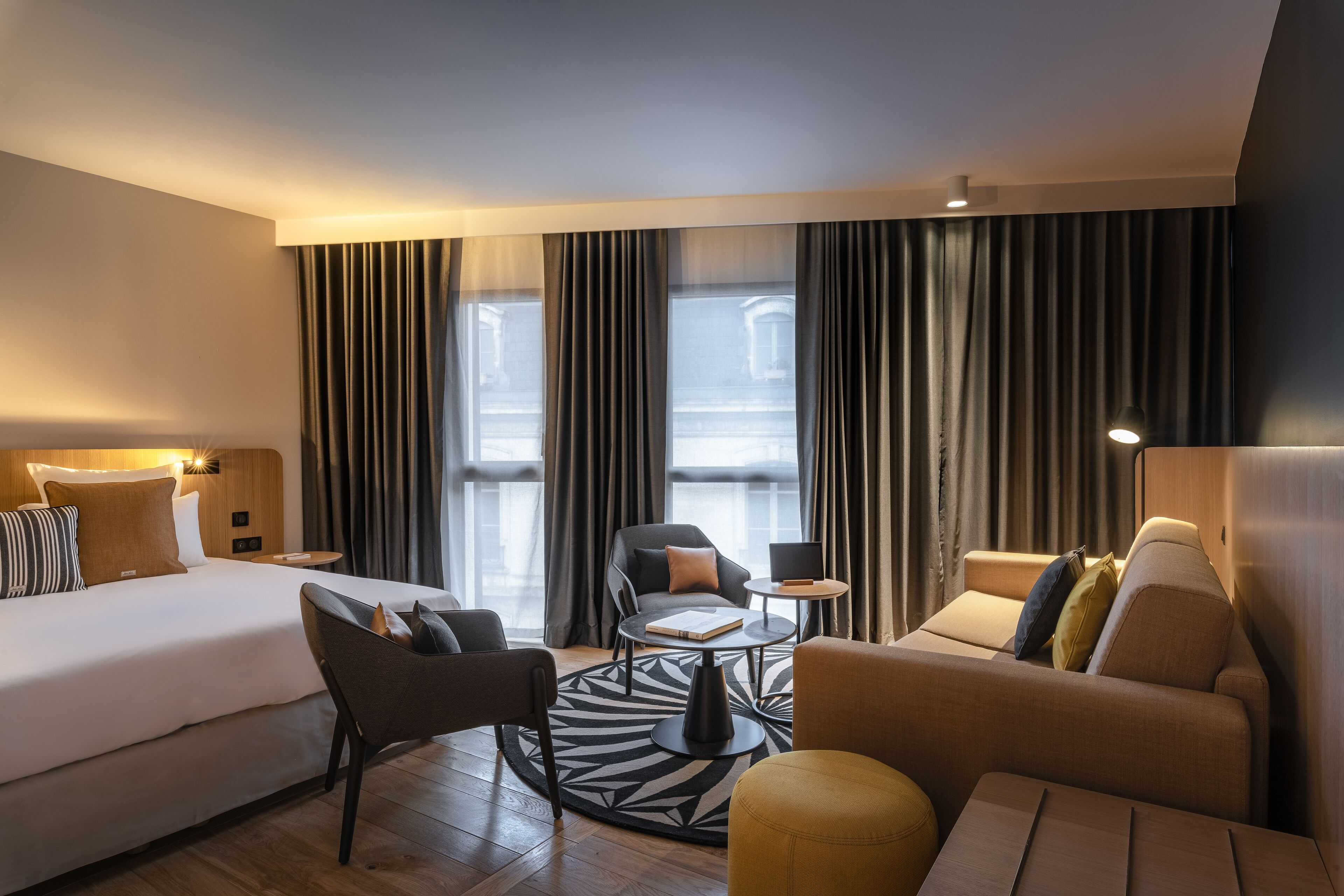 Habitación de hotel moderna con cama, sofá, sillón, una mesita y vistas de la ciudad a través de amplias ventanas.