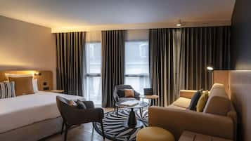 Habitación de hotel moderna con cama, sofá, sillón, una mesita y vistas de la ciudad a través de amplias ventanas.