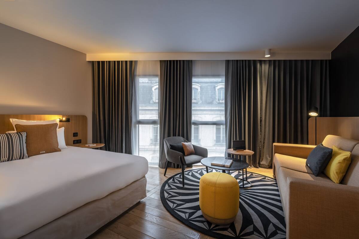 Une chambre d’hôtel moderne, dotée d’un grand lit, d’un canapé, d’un fauteuil, d’une petite table et offrant une vue sur la ville grâce à de grandes fenêtres.