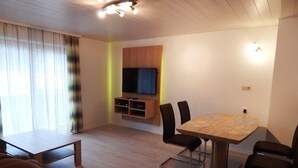 Een moderne woonkamer met een houten eettafel, zwarte stoelen, een bank, een aan de muur bevestigde televisie en een groot raam met lichte gordijnen.