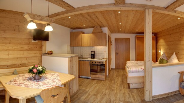 Um quarto acolhedor em estilo cabana, com mesa de jantar de madeira, uma cadeira, cama com almofadas, pequena cozinha com fogão e pia, e teto de madeira.