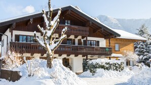 Um chalé de madeira de dois andares com varandas, cercado por terreno e árvores cobertas de neve.