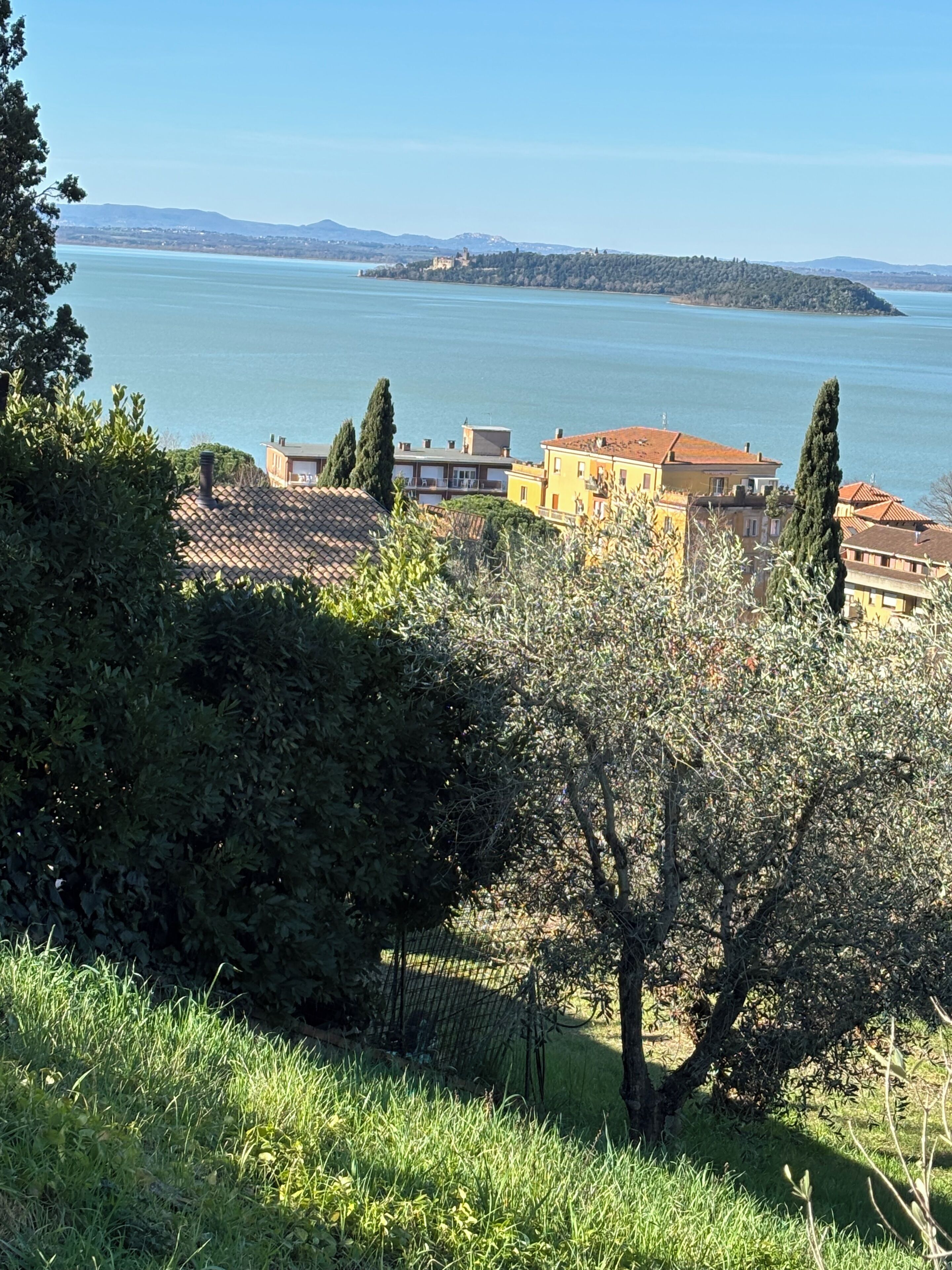 A coastal town with buildings near a body of water, a clear sky, and a hilly landscape in the distance.