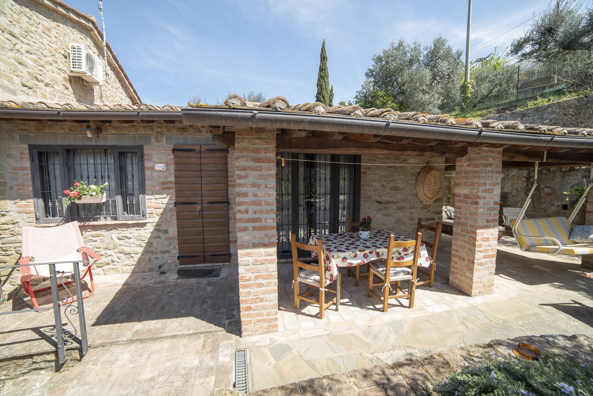 A rustic stone building with a tiled roof, a patio with a table and chairs, and a small garden area.