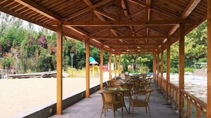 Un espace extérieur aménagé, abrité et doté de tables et de chaises en bois, entouré d’une balustrade en bois, offrant une vue sur une plage de sable et une végétation luxuriante.