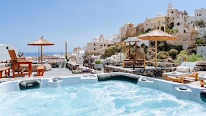 Un área de piscina en la azotea con jacuzzis, sillones de descanso y sombrillas, con vistas a una ciudad costera de edificios blancos y cielo despejado.