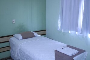 Una cama individual con colchón blanco y almohada marrón, una mesita con una toalla blanca doblada, y una ventana con cortinas blancas.