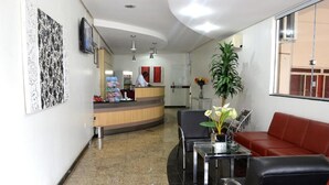 A reception area with a counter, a red sofa, a glass table with a vase, and a potted plant. - Domus Hotel Veneza (Ipatinga)