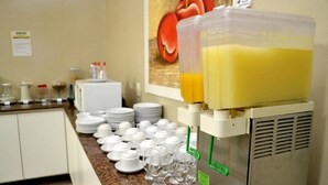 A juice dispenser with a large container, a refrigerator, and a counter with stacked plates and glasses. - Domus Hotel Veneza (Ipatinga)