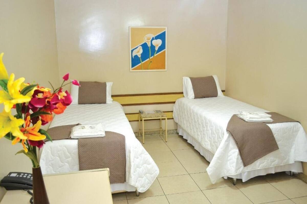 A hotel room with two beds, a small table, a vase with flowers, and a painting on the wall.